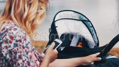 母亲摇摆婴儿车和使用智能手机在湖边。 夏日阳光明媚。
